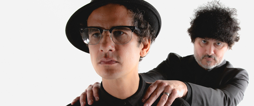 Portrait of two men, one wearing a hat and the second one is grabbing his shoulder.
