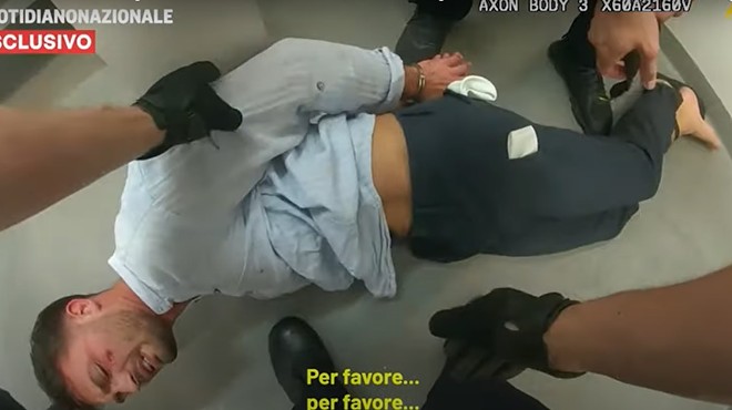 A student is lying face down on the floor of a holding cell, with his handcuffed hands behind his back