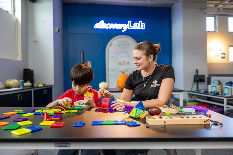 Image: A child and a woman play with puzzles and toys on a table. A neon sign behind them reads, "discoverLab."