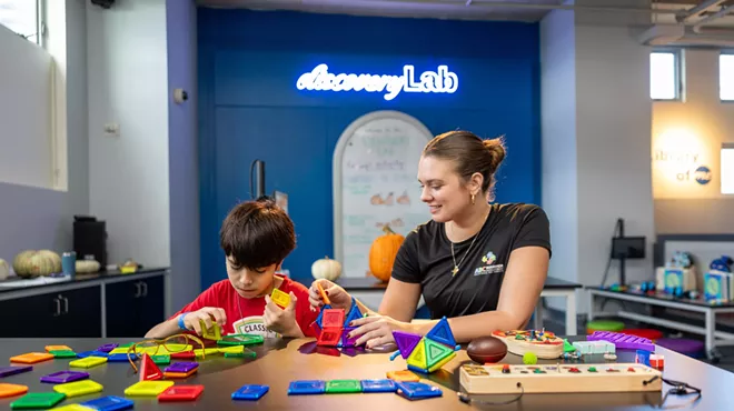 A child and a woman play with puzzles and toys on a table. A neon sign behind them reads, "discoverLab."