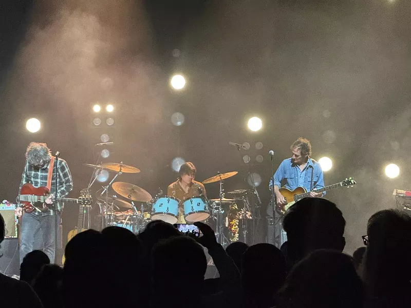 Image: A rock band on stage, rocking, with the silhouettes of audience members in the foreground