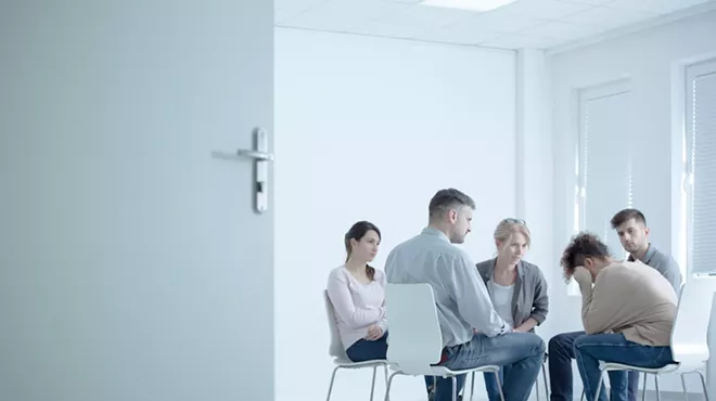 stark stock photo with much white space showing a woman crying in a small seated group at a rehab center with an open door in the foreground